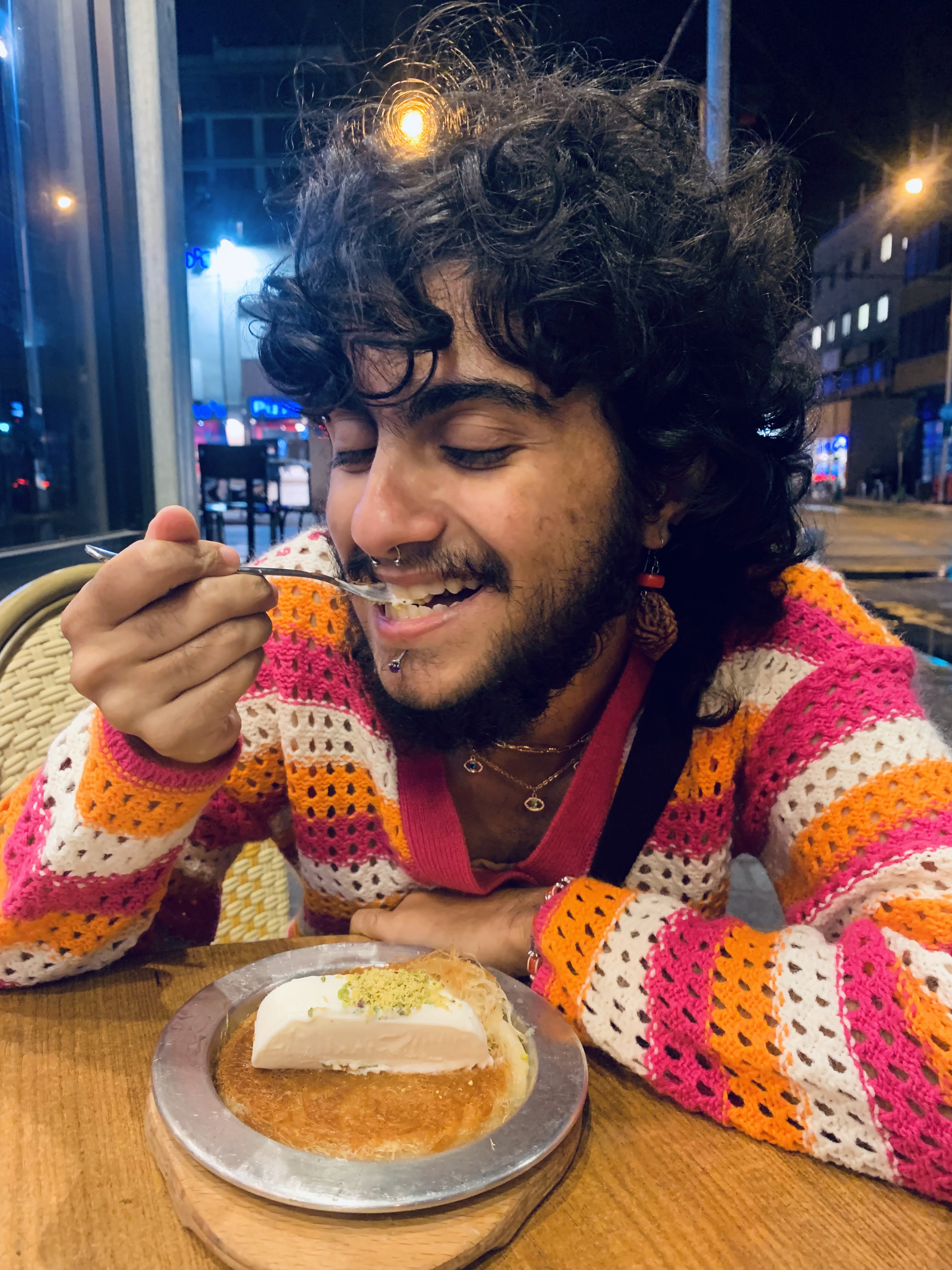 Mel with black curly hair wearing a colorful knitted sweater and jewellery smiles while sitting in front of and eating a spoonful of Knafeh and Turkish Ice Cream with crushed pistachio on top.