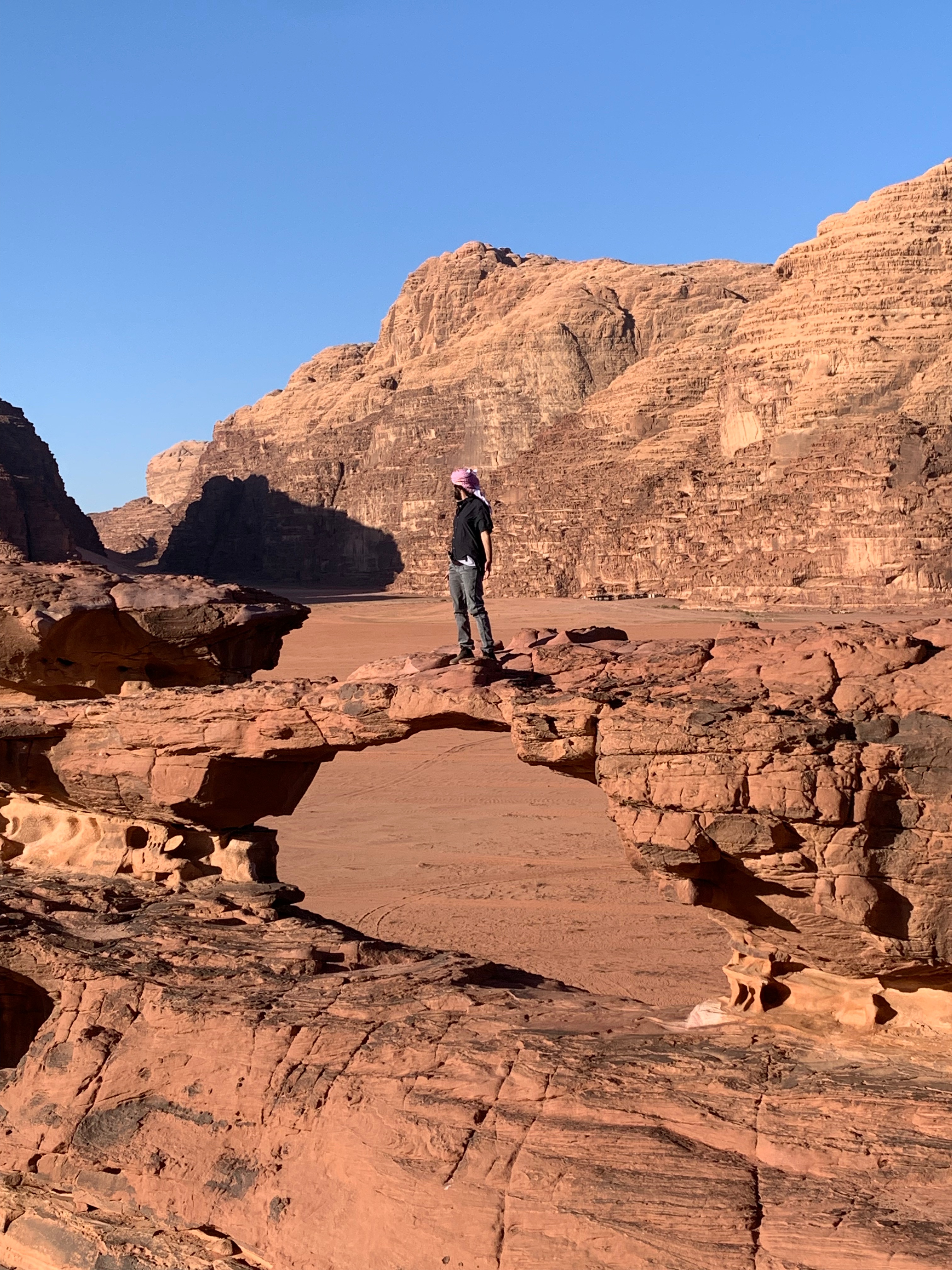 Mel in grey jeans, black shirt and red keffiyeh standing on a natural rock arch in a desert landscape with steep, rocky cliffs in the background under a clear blue sky.